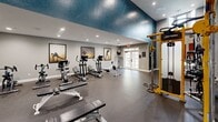 Located at 5720 Forest Park Rd in Dallas, TX, this fitness center offers a spacious and well-appointed exercise environment designed to support a wide range of workout routines. The room is defined by its dramatically soaring double-height ceilings, which lend an open and airy atmosphere rarely found in a gym of this footprint. Large arched transom windows and French doors line the walls, inviting abundant natural light while providing views to the exterior. The interior walls are painted in a cool gray palette, accented by a bold deep-blue textured ceiling detail that adds visual character to the space. Durable rubber flooring covers the entire area, providing both safety and functionality. The fitness floor is comprehensively equipped with treadmills, ellipticals, a rowing machine, and stationary bikes for cardiovascular training, alongside a yellow Life Fitness functional cable trainer with TRX and battle rope attachments for functional and strength workouts. A full dumbbell rack with adjustable benches and a wall-mounted mirror round out the strength training options. Motivational athletic artwork decorates the walls, and posted fitness center rules ensure a safe environment for all users. Recessed lighting supplements the natural light, keeping the space bright and energizing throughout the day.
