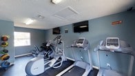 This fitness center offers a focused and functional environment for residents to maintain an active lifestyle. The space is outfitted with a variety of cardio equipment, including treadmills, an elliptical trainer, and a Precor cross-trainer, providing ample options for cardiovascular workouts. A dedicated free weights area with a dumbbell rack rounds out the strength training offerings. Two wall-mounted flat-screen televisions are positioned for easy viewing from the cardio machines, allowing users to stay entertained during their workouts. The room features cool-toned blue-gray walls that create a calm, motivating atmosphere, complemented by a textured ceiling with integrated lighting. Dark flooring anchors the space and provides a durable surface suited for exercise. A window allows for natural light to enter, adding to the overall brightness of the room. The layout is thoughtfully organized to maximize the available floor space while keeping equipment accessible and easy to navigate. This fitness center delivers a convenient, amenity-rich workout facility all on a single level.