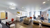 This sophisticated single-floor space showcases a contemporary open-concept design that seamlessly blends living, dining, and kitchen areas into one cohesive environment. The kitchen features a large white island with dark bar seating, complemented by clean cabinetry and modern finishes throughout. An eye-catching sculptural chandelier anchors the living area, adding an artistic focal point to the bright white ceiling above. The living space is thoughtfully furnished with a neutral-toned sectional sofa, accented by bold pops of red and orange through decorative pillows and accent chairs, creating a dynamic yet balanced aesthetic. Floor-to-ceiling windows line the exterior wall, flooding the interior with generous natural light and offering expansive views of the surrounding cityscape. The open layout is further defined by a large geometric-patterned area rug that grounds the seating arrangement. Throughout the space, a consistent palette of white, grey, and neutral tones is elevated by carefully chosen accent pieces, lending the interior a polished, upscale character. The overall ambiance is one of modern elegance — bright, airy, and meticulously designed for both comfort and style.