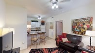 This single-floor residence features an open-concept design that integrates the living, dining, and kitchen areas into one cohesive and functional space. The living area is anchored by plush carpeting and a bold floral-patterned area rug, complemented by a comfortable dark leather sofa accented with red and gold throw pillows. A ceiling fan with a light fixture overhead provides both comfort and illumination throughout the space.

The kitchen is efficiently laid out with white cabinetry extending to the ceiling, offering generous storage capacity. White appliances, including a refrigerator and microwave, contribute to a clean, uniform aesthetic. Dark countertops provide a subtle contrast to the light cabinetry. A small dining nook with a bistro-style table and two decorative chairs sits adjacent to the kitchen, making it an ideal spot for casual meals.

A hallway visible from the main living area leads to additional rooms, with doors providing privacy and separation. The overall color palette of whites, creams, and warm neutrals creates a bright, welcoming ambiance throughout the space, while decorative accents add personality and warmth.