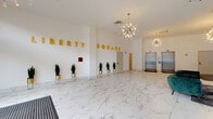 Liberty Square presents a grand, single-floor lobby that immediately commands attention with its sophisticated design and upscale finishes. The expansive entrance area is anchored by large-format white marble-style porcelain tile flooring, with subtle grey veining that runs throughout the entire space, lending an air of elegance and continuity.

The lobby's focal wall features bold, three-dimensional gold lettering spelling out "Liberty Square," serving as a striking visual centerpiece. A row of tall snake plants in gold geometric stands lines this wall, adding a touch of greenery that contrasts beautifully against the bright white interior.

Overhead, a pair of mid-century modern sputnik chandeliers cast a warm, ambient glow across the space, contributing to the overall contemporary aesthetic. High ceilings amplify the sense of openness and volume throughout.

A dedicated lounge area is furnished with deep emerald green velvet sofas and accent chairs arranged around glass-top coffee tables, offering a comfortable and stylish setting for residents or guests. Floor-to-ceiling windows along the lounge perimeter flood this zone with abundant natural light and provide views of the surrounding streetscape.

Two stainless steel elevators are conveniently positioned at the rear of the lobby, providing access to upper floors. The overall ambiance is clean, modern, and welcoming — a polished gateway that sets a high standard for the building it represents.