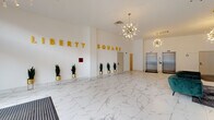 Liberty Square presents a grand, single-floor lobby that immediately commands attention with its sophisticated design and upscale finishes. The expansive entrance area is anchored by large-format white marble-style porcelain tile flooring, with subtle grey veining that runs throughout the entire space, lending an air of elegance and continuity.

The lobby's focal wall features bold, three-dimensional gold lettering spelling out "Liberty Square," serving as a striking visual centerpiece. A row of tall snake plants in gold geometric stands lines this wall, adding a touch of greenery that contrasts beautifully against the bright white interior.

Overhead, a pair of mid-century modern sputnik chandeliers cast a warm, ambient glow across the space, contributing to the overall contemporary aesthetic. High ceilings amplify the sense of openness and volume throughout.

A dedicated lounge area is furnished with deep emerald green velvet sofas and accent chairs arranged around glass-top coffee tables, offering a comfortable and stylish setting for residents or guests. Floor-to-ceiling windows along the lounge perimeter flood this zone with abundant natural light and provide views of the surrounding streetscape.

Two stainless steel elevators are conveniently positioned at the rear of the lobby, providing access to upper floors. The overall ambiance is clean, modern, and welcoming — a polished gateway that sets a high standard for the building it represents.