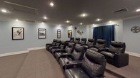 Located at 3150 Skinner Mill Rd in Augusta, Georgia, this dedicated home theater room delivers a true cinematic experience within a residential setting. The space is finished with soft blue-gray walls, white crown molding, and recessed lighting set against a darker ceiling, creating an atmosphere reminiscent of a private screening room. Wall-to-wall carpet in a neutral tone adds comfort underfoot and contributes to the room's acoustics.

The seating arrangement consists of multiple rows of black leather recliner chairs equipped with built-in cup holders, providing ample accommodation for family and guests alike. A large flat-screen television is mounted on the front wall above a wooden media console with a granite-style top, which houses an organized collection of movies, games, and media equipment, flanked by floor-standing speakers for enhanced audio.

Cinema-themed décor lends character throughout the space, including framed movie posters—such as a prominent Rocky print—film reel wall art, and a wall-mounted media rack. A classic popcorn machine stationed in the corner adds a fun, authentic touch. Deep blue curtains along one wall complete the theater ambiance. Crown molding and recessed lighting throughout lend a polished, well-appointed finish to this thoughtfully designed entertainment space.