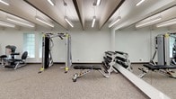 This fitness center offers a comprehensive workout environment designed to accommodate a range of exercise routines. The space is finished with neutral-toned carpet flooring and light gray walls, creating a clean and functional atmosphere throughout. Distinctive exposed beam detailing runs across the ceiling, complemented by rows of bright linear fluorescent fixtures that ensure thorough, even illumination across the entire floor.

The cardio zone is well-appointed with multiple treadmills and an elliptical machine, each positioned facing wall-mounted flat-screen televisions for an engaging workout experience. A large mirror along one wall visually expands the space and allows users to monitor their form. The strength training area features a multi-function cable machine, an adjustable weight bench, and a tiered dumbbell rack stocked with a full range of free weights.

Additional equipment includes a leg extension machine situated near glass block windows that allow natural light to filter in. A dedicated refreshment station along one wall includes dark cabinetry with a granite-style countertop, a water cooler, and a small kitchen area equipped with a full-size refrigerator and microwave, ensuring convenience during workouts.