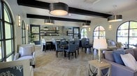 This single-floor space exudes a contemporary yet inviting ambiance, thoughtfully designed for dining and socializing. The room is anchored by striking exposed dark beams that contrast beautifully against the white ceiling, adding architectural depth and character. Underfoot, a herringbone-patterned stone tile floor lends a refined, textural element that ties the space together cohesively.

Multiple dining groupings are arranged throughout, featuring dark wood tables paired with upholstered charcoal chairs, offering ample seating for gatherings. A standout feature wall showcases stacked stone tile cladding, complemented by floating shelves styled with decorative greenery and curated accents, creating a warm focal point for the room.

Industrial-style drum pendant lights hang at varying heights, providing both functional illumination and bold visual interest. Arched windows along one wall invite natural light and offer glimpses of the outdoors, softening the modern aesthetic with a classic architectural touch. Framed botanical artwork and lush potted plants are positioned throughout, adding organic warmth. The overall design balances modern sophistication with comfortable, welcoming details.