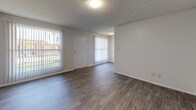 This single-floor residence offers a practical and straightforward layout designed for comfortable everyday living. The living area serves as the central hub of the unit, featuring durable wood-style flooring, neutral white walls, and a textured ceiling. Large windows fitted with vertical blinds allow natural light to fill the space, with views of an outdoor green area visible from the front entry.

The galley-style kitchen is efficiently arranged, equipped with a gas range, full-size refrigerator, double-basin sink, and ample upper and lower cabinetry in a warm wood tone. Tile backsplash lines the cooking area, and the countertops provide functional workspace throughout.

The bedrooms are carpeted and share a consistent design element: painted white brick accent walls that add a subtle textural character to each room. Each bedroom includes vertical-blind-covered windows that introduce natural light, and the textured ceilings carry throughout. One room features wood-style flooring instead of carpet, offering added flexibility.

The bathroom includes a full tub and shower combination with white tile surround, a wood-toned vanity with a stone-look countertop, and chrome fixtures. Wood-style flooring completes the space. Overall, the unit presents a clean, functional environment with consistent finishes throughout.