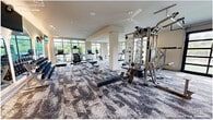 This expansive fitness center offers a comprehensive, amenity-rich workout environment spread across a single open floor. The main workout area features patterned carpet flooring, structural columns, and generous ceiling height, creating a bright and airy atmosphere. Large floor-to-ceiling windows and a glass-panel garage door flood the space with natural light and provide views of the surrounding outdoor greenery. The main floor is fully equipped with a wide variety of strength-training machines, cable systems, free-weight benches, a full dumbbell rack, and multiple cardio machines including treadmills. Ceiling fans are installed throughout to maintain comfort during workouts. A dedicated studio room offers a distinct aesthetic with a deep navy ceiling, arched mirrored wall accents, warm accent lighting, and light hardwood flooring — ideal for yoga, stretching, or group fitness sessions. Back in the main area, arched mirror panels with integrated backlighting add an upscale design element, while wall-mounted TVs provide entertainment. A functional area also features a heavy bag and a multi-use rig for functional fitness training. Barn-style sliding doors lead to a well-appointed restroom or lounge area, completing this thoughtfully designed, full-service fitness facility.
