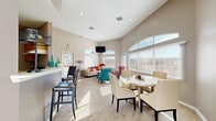 This single-floor community space is thoughtfully designed to foster gathering and relaxation, offering an open-plan layout that seamlessly connects multiple functional zones. Vaulted ceilings rise throughout the main area, amplifying the sense of openness and volume, while a series of arched transom windows and large blinded windows flood the interior with generous natural light.

The lounge area is anchored by a patterned area rug and furnished with a neutral sofa accented by colorful throw pillows, a vibrant patterned wingback chair, and a round coffee table — all centered beneath a wall-mounted flat-screen television. Adjacent to the lounge, a dedicated dining area features a round table with cream upholstered chairs, ideal for group gatherings. Light wood-style flooring runs throughout these areas, lending warmth and continuity.

The kitchenette is defined by a raised bar counter with leather bar stools, wooden cabinetry, and a refrigerator, providing a practical amenity hub for the space. Decorative accents including abstract wall art and teal glass vases add personality and color throughout.

A secondary tiled area with recessed lighting and high ceilings offers additional flexible space. The overall ambiance is welcoming and contemporary, characterized by neutral warm-grey tones, ample daylight, and a balanced mix of comfort and functionality.