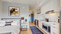 This thoughtfully designed studio unit offers a bright, open-concept layout that efficiently combines living, sleeping, and dining areas into one cohesive space. Light hardwood flooring flows throughout, anchoring the clean, contemporary aesthetic defined by neutral tones and modern finishes.

The sleeping area features a full-size bed frame with a muted accent wall adorned with bold artwork, adding a touch of character. Large windows along the far wall flood the living area with natural light while framing impressive elevated views of the city skyline and surrounding treetops — a standout feature of this unit.

The kitchen is well-equipped with flat-front cabinetry in a warm wood tone, open shelving, stainless steel appliances including a full-size refrigerator, a built-in oven, and an induction cooktop. A compact dining nook with a desk and chair provides a practical workspace or casual dining area.

A dedicated in-unit laundry area with a stacked washer and dryer is seamlessly integrated into the layout near the entry, adding everyday convenience. The bathroom features hardwood-style flooring, a clean white vanity, and a shower-tub combination. The entry corridor, finished with a teal accent door, leads smoothly into the main living space, completing this efficient and stylish unit.