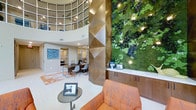 This single-floor space makes an immediate impression through its architectural grandeur and thoughtful design details. The interior rises dramatically with a curved, multi-paned clerestory ceiling that floods the space with natural light, creating an airy, open atmosphere despite its intimate scale. A commanding living green wall serves as a focal centerpiece, showcasing a lush arrangement of preserved moss, foliage, and flowering plants accented by integrated spotlights, bringing a natural, biophilic element into the refined interior. Adjacent to the green wall, a sleek reception or service counter with dark cabinetry and a white surface provides a functional workspace. A geometrically faceted column clad in warm metallic bronze tones adds a sculptural contrast to the organic textures nearby. The seating area features a richly upholstered burnt-orange sofa paired with a minimalist side table, offering a comfortable lounge space. Glass-partitioned offices in the background allow for visual connectivity while maintaining privacy. Contemporary artwork, patterned area rugs, and carefully curated furnishings complete the polished, professional ambiance throughout.