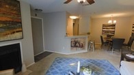 This single-floor residence showcases an open-concept design that seamlessly connects the living, dining, and kitchen areas. The living space is anchored by a patterned area rug and a ceiling fan that provides both function and visual interest overhead. A half-wall breakfast bar serves as the dividing element between the kitchen and the common living areas, topped with a pendant light and accented by decorative signage on its face, adding a rustic, personalized touch. The kitchen, visible beyond the bar, features light-toned cabinetry and a refrigerator, with warm overhead lighting creating a welcoming atmosphere. Adjacent to the kitchen, a dining area accommodates a table and chairs, including metal bar-style seating that complements the casual aesthetic of the space. The entryway, visible to the left, features a closet with a paneled door and transitions from hard flooring to the carpeted living area. Neutral gray walls throughout provide a versatile backdrop, while the layout promotes a sense of openness and easy flow between functional zones. The overall ambiance is casual and comfortable, with decor details adding character to the space.
