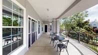This single-level outdoor space features a generously proportioned covered corridor and patio area, thoughtfully designed to blend functionality with aesthetic appeal. The walkway runs the length of the building, framed by sturdy decorative columns finished in neutral gray tones with crisp white trim detailing — a classic architectural touch that lends the space a polished, welcoming character.

Multiple entry doors with glass panels line the interior wall, each accented with bold purple finishes and white surrounds that add a distinctive visual contrast. Large multi-pane windows allow natural light to permeate the corridor throughout the day.

The open-air side of the walkway is enclosed by ornamental iron railings, offering an unobstructed view of the lush, tree-lined surroundings, including mature palms, flowering shrubs, and neighboring rooftops. The verdant outdoor scenery creates a serene, park-like ambiance visible from the patio area.

Seating is thoughtfully arranged along the corridor, including round bistro tables with cross-back chairs and a classic wooden bench — ideal for relaxing or socializing. A branded GWR Management door mat marks the main entry point, reinforcing the professionally managed nature of the property.

The smooth concrete flooring throughout is clean and durable, completing a well-maintained outdoor space that offers both comfort and connectivity to the surrounding landscape.