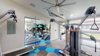 This single-level fitness center is thoughtfully designed to support a wide range of workout styles in a bright, open environment. The space is divided into distinct functional zones, each optimized for a specific type of training. A cardio area lines one wall, equipped with multiple treadmills positioned in front of expansive glass garage-style doors that flood the room with natural light and offer views of the surrounding greenery. Adjacent to this, a strength and cable machine zone houses multi-function cable systems and resistance equipment, with ceiling-mounted pulley systems maximizing vertical space. A dedicated cycling area features stationary spin bikes beneath large clerestory windows that frame the tree canopy outside. The free weights and functional training section is well-stocked with dumbbells, stability balls, resistance bands, step platforms, and specialty training machines. Full-length mirrored walls run throughout the space, enhancing the sense of openness and allowing users to monitor form. The interior is finished with a striking hexagonal tile floor in shades of blue, teal, and gray, adding a bold visual identity to the space. Oversized ceiling fans maintain airflow, and recessed lighting ensures consistent illumination. The overall ambiance is modern, energetic, and purpose-built for a comprehensive fitness experience.