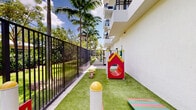 Located at 21 NE First Ct in Dania Beach, Florida, this well-appointed outdoor amenity space offers a variety of recreational features set against a backdrop of lush South Florida landscaping. The grounds are thoughtfully designed to serve multiple user groups, blending green space with dedicated activity zones throughout.

A standout feature is the children's play area, enclosed by a sturdy black metal fence for safety. This colorful zone is surfaced with artificial turf and equipped with a red-and-yellow playhouse structure, a tunnel crawl-through, and vibrant stepping stools, providing a fun and secure environment for younger visitors.

Adjacent to the play area, a concrete walkway winds through the property, lined with an outdoor fitness circuit. Green exercise stations — including a rowing machine, body resistance equipment, and stretching apparatus — are anchored along the path, each accompanied by instructional signage, making fitness accessible in an open-air setting.

The landscaping throughout is mature and tropical, with tall royal palms, broad-canopy trees, and dense shrub borders framing the perimeter. Well-maintained grass lawns, mulched garden beds, and the surrounding wrought-iron fencing contribute to a sense of privacy and order. The space benefits from abundant natural sunlight and open sky views, creating an inviting outdoor environment reflective of the vibrant South Florida lifestyle.