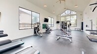 Located at 2400 N Morrison Blvd in Hammond, Louisiana, this fitness center is a well-appointed, single-floor exercise facility designed to support a complete workout experience. The space is immediately distinguished by its dramatic vaulted ceiling, which rises to a striking peak and is fitted with dual ceiling fans to maintain airflow throughout. Large floor-to-ceiling windows and wide panel windows line multiple walls, flooding the interior with natural light while offering open views of the surrounding tree-lined landscape and outdoor amenity areas. The durable rubber-coated flooring provides a practical and resilient surface throughout. Equipment is thoughtfully arranged to maximize usable space, featuring multiple treadmills positioned directly in front of the expansive windows, a multi-station cable and weight machine, a free-weight dumbbell rack, adjustable benches, and additional strength training stations. Wall-mounted flat-screen televisions are positioned at key viewing angles across the room, and a large mirror along one wall enhances the sense of space while allowing users to monitor their form. Pendant lighting supplements the abundant natural light for evening use. The overall ambiance is clean, bright, and functional, creating an inviting environment well-suited for daily fitness routines.