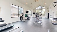 Located at 2400 N Morrison Blvd in Hammond, Louisiana, this fitness center is a well-appointed, single-floor exercise facility designed to support a complete workout experience. The space is immediately distinguished by its dramatic vaulted ceiling, which rises to a striking peak and is fitted with dual ceiling fans to maintain airflow throughout. Large floor-to-ceiling windows and wide panel windows line multiple walls, flooding the interior with natural light while offering open views of the surrounding tree-lined landscape and outdoor amenity areas. The durable rubber-coated flooring provides a practical and resilient surface throughout. Equipment is thoughtfully arranged to maximize usable space, featuring multiple treadmills positioned directly in front of the expansive windows, a multi-station cable and weight machine, a free-weight dumbbell rack, adjustable benches, and additional strength training stations. Wall-mounted flat-screen televisions are positioned at key viewing angles across the room, and a large mirror along one wall enhances the sense of space while allowing users to monitor their form. Pendant lighting supplements the abundant natural light for evening use. The overall ambiance is clean, bright, and functional, creating an inviting environment well-suited for daily fitness routines.