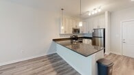 Located at 4201 Runner Loop in Chester, Virginia, this single-floor residence offers a thoughtfully designed layout with modern finishes throughout. The kitchen serves as a central highlight, featuring white shaker-style cabinetry extending to the ceiling, dark granite countertops, and a full suite of stainless steel appliances including a range, built-in microwave, and refrigerator. A peninsula with an undermount sink and pendant lighting above creates a natural gathering point and provides a seamless connection to the adjacent living area. Light wood-style flooring runs throughout the main living spaces, contributing to a clean, cohesive aesthetic. The bedroom is carpeted for comfort and includes a ceiling fan, as well as windows overlooking a lush, green outdoor setting with access to a balcony railing visible beyond. A conveniently located in-unit laundry closet with stacked washer and dryer is accessible from the bedroom area. The full bathroom is well-equipped with a vanity topped in dark granite, white cabinetry with black hardware, a mirror, and a tub-shower combination with white tile surround. Neutral wall tones and consistent finishes throughout create a bright, move-in-ready environment.