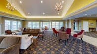 Located at 7113 Eastern Woods Pkwy in Findlay, Ohio, this single-floor community clubhouse offers a refined and inviting atmosphere designed for social gatherings and everyday comfort. The grand entrance foyer greets visitors with an ornate patterned carpet, recessed lighting, crown molding, and warm gold-toned walls that carry throughout the space, establishing a cohesive and elegant design language.

The expansive main lounge serves as the heart of the facility, featuring a dramatic tray ceiling with a chandelier, large windows along multiple walls that flood the room with natural light, and comfortable seating arrangements including sofas and accent chairs arranged to encourage conversation. An adjoining sunlit nook with a bay window provides a casual dining area, ideal for smaller gatherings.

The formal dining room accommodates large groups around a substantial wooden table, complemented by pendant lighting, a tray ceiling, and generous windows overlooking the surrounding green landscape. A fully equipped kitchen supports the dining areas with rich cherry cabinetry, stainless steel appliances, granite countertops, tile flooring, and a tray ceiling with recessed lighting. Additional amenities visible throughout include a game area and open corridors connecting the various spaces seamlessly, making this clubhouse a versatile and welcoming hub for its community.