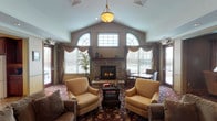 This single-floor community space is thoughtfully designed to foster comfort and connection. The centerpiece is a grand great room with soaring vaulted ceilings and a striking floor-to-ceiling stone fireplace, flanked by elegantly draped arched windows that fill the space with natural light. A pendant light fixture hangs above multiple seating arrangements, including plush armchairs and sofas set atop rich, floral-patterned carpeting that flows throughout the entire space.

The layout opens generously into additional lounge areas, where residents can gather around a flat-screen television, settle into cozy seating nooks, or make use of a dedicated dining area furnished with wooden tables and upholstered chairs. Framed artwork and carefully chosen accent pieces lend a warm, residential character to each zone.

A pass-through kitchen features rich cherry wood cabinetry, stone-tile flooring, ample counter space, and full appliances including a range, built-in microwave, and dishwasher — well-suited for entertaining or catered events.

The space is accessed through a set of classic wood-and-glass double doors, flanked by sidelights that preview the bright, inviting interior. Throughout, the design balances traditional elegance with practical functionality, creating an atmosphere that feels both refined and approachable.