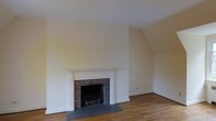 This single-floor space exudes a timeless, classic charm through its carefully considered architectural details and clean, understated finishes. The living area is anchored by a traditional wood-burning brick fireplace, framed by an elegant white painted mantel with fluted pilaster columns and a flat entablature, lending the room a sense of refined character. A slate hearth extends outward from the firebox, providing both a functional and aesthetic grounding element. The walls are finished in a soft, neutral cream tone that reflects light evenly throughout the space, contributing to an open and airy ambiance. Warm-toned hardwood flooring runs throughout, adding natural texture and continuity underfoot. The room benefits from clean white baseboards and trim work that complement the overall classic aesthetic. The ceiling is smooth and generously proportioned, enhancing the sense of volume within the space. A subtle angled wall detail in one corner suggests an architectural transition, possibly to an adjoining area or stairwell, adding a layer of spatial interest. Overall, the space presents a well-maintained, traditional interior well-suited to a variety of uses.
