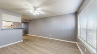 This single-floor, 1-bedroom, 1-bathroom unit offers a practical and comfortable layout designed to make efficient use of its space. The combined kitchen and living area forms the heart of the unit, where light wood-style laminate flooring and neutral gray walls create a clean, cohesive aesthetic. The living room is equipped with a ceiling fan and light fixture, providing both comfort and illumination. A pass-through counter connects the kitchen to the living area, offering an open feel between the two spaces. The kitchen features white cabinetry, dark granite-style countertops, white appliances including a refrigerator, dishwasher, and electric range, along with overhead fluorescent lighting. The spacious bedroom includes a ceiling fan, a window with blinds that allows natural light, and the same light wood-style flooring found throughout the unit. The bathroom is appointed with a single-sink vanity featuring dark countertops, a full tub-shower combo with white tile surround, and a large mirror. A hallway connects the main living spaces to the bedroom and bathroom. Textured ceilings and white baseboards are consistent throughout, contributing to the unit's tidy, move-in-ready presentation.