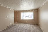 This single-floor unit offers a practical and straightforward layout across several distinct rooms. The living area is generously proportioned, finished with neutral-toned carpet and warm beige walls. A decorative border trim runs along the upper perimeter, adding a subtle design detail, while vertical blinds on a large window allow natural light to filter in. The textured ceiling is consistent throughout the carpeted areas of the unit.

The kitchen is equipped with white cabinetry, both upper and lower, paired with a continuous countertop featuring an integrated stainless steel sink. A gas range with overhead ventilation hood anchors one wall, while a multicolored mosaic tile backsplash in neutral earth tones provides visual interest throughout the cooking area.

The bedrooms are carpeted and feature neutral walls, window blinds for privacy, and textured ceilings. At least one bedroom includes a built-in closet with bifold doors and a rod for hanging storage. Each room benefits from natural light through exterior-facing windows.

The bathroom features classic pink ceramic wall tiles with a white accent border, a mosaic tile floor, and a full soaking tub. A textured ceiling completes the space, which connects directly to the hallway via a dark-framed door.