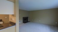 This single-floor space presents a straightforward and functional layout anchored by a spacious living area. The room is finished with wall-to-wall carpeting in a neutral tone, contributing to a quiet and comfortable atmosphere underfoot. The walls are painted in an earthy olive-green hue, lending warmth to the interior while maintaining a understated, versatile aesthetic.

A dark-mantled fireplace serves as the focal point of the living area, providing both visual interest and a practical source of warmth. The fireplace surround features a simple, clean design with a dark hearth extending onto the carpeted floor. The ceiling is finished with a textured surface and painted a crisp white, helping to brighten the space and enhance the sense of openness.

A partial view of an adjacent area — likely a kitchen or utility space — is visible along the left side, suggesting a connected, open-concept flow between living and functional zones. The space includes standard electrical outlets positioned at regular intervals along the walls, as well as a wall-mounted utility panel near the fireplace. Overall, the space offers a clean, move-in-ready foundation with classic residential finishes.