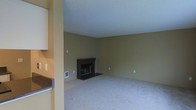 This single-floor space presents a straightforward and functional layout anchored by a spacious living area. The room is finished with wall-to-wall carpeting in a neutral tone, contributing to a quiet and comfortable atmosphere underfoot. The walls are painted in an earthy olive-green hue, lending warmth to the interior while maintaining a understated, versatile aesthetic.

A dark-mantled fireplace serves as the focal point of the living area, providing both visual interest and a practical source of warmth. The fireplace surround features a simple, clean design with a dark hearth extending onto the carpeted floor. The ceiling is finished with a textured surface and painted a crisp white, helping to brighten the space and enhance the sense of openness.

A partial view of an adjacent area — likely a kitchen or utility space — is visible along the left side, suggesting a connected, open-concept flow between living and functional zones. The space includes standard electrical outlets positioned at regular intervals along the walls, as well as a wall-mounted utility panel near the fireplace. Overall, the space offers a clean, move-in-ready foundation with classic residential finishes.