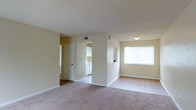 This single-floor residence offers a straightforward and functional layout designed for comfortable everyday living. The main living area is finished with wall-to-wall carpeting in a neutral tone, providing a soft underfoot feel throughout the space. A large window along one wall allows natural light to filter into the room, brightening the interior and creating an airy atmosphere. The walls are painted in a warm beige, contributing to a clean and cohesive look.

A central structural column partially divides the living area from the adjacent spaces, adding subtle definition without fully closing off the floor plan. From the living area, there is a clear sightline into the kitchen, which features white cabinetry and appears to have hard flooring, contrasting with the carpeted main area. HVAC vents are positioned in the ceiling, indicating a centralized climate control system throughout the unit.

A hallway visible from the main living space leads to additional rooms, with a white door providing access to what appears to be a secondary area or utility space. The overall ambiance is understated and move-in ready, with a neutral palette that offers flexibility for personalization.