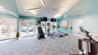 Located at 1000 Forest Oak Drive in Charlotte, NC, this fitness center offers a thoughtfully designed exercise space for residents. The room is characterized by soaring vaulted ceilings that create an open, airy atmosphere, complemented by vibrant teal walls and soft patterned carpeting throughout. Modern linear pendant lighting and a ceiling fan ensure the space remains bright and comfortable during any workout.

The cardio zone features multiple treadmills, an elliptical trainer, and a stair climber, each equipped with mounted flat-screen monitors for entertainment. A dedicated strength training area includes a cable machine, a leg press, a stationary spin bike, a weight bench, and a full dumbbell rack with additional functional training accessories. Mirrors along the walls enhance the sense of space and allow for form checks during exercise.

Large windows and glass doors flood the interior with abundant natural light while offering views of the surrounding trees and outdoor spaces. French doors provide direct access to an outdoor patio and pool area, seamlessly blending indoor and outdoor amenities. A colorful framed art piece adds a decorative touch to the space. The fitness center is accessible via a short staircase entry and is well-suited to support a comprehensive daily workout routine.
