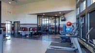 This fitness center is thoughtfully designed to accommodate a wide variety of exercise needs within a single, open-plan floor. The space features a clean, modern aesthetic with neutral walls accented by a bold dark red horizontal stripe that adds visual structure and energy to the environment. Recessed ceiling lighting ensures the room is well-lit throughout, while a wall-mounted flat-screen TV provides entertainment during workouts. A row of treadmills and cardio machines lines one side of the space, offering dedicated areas for cardiovascular training. On the opposite end, a dumbbell and medicine ball rack — stocked with an array of colorful exercise balls — provides ample free weight options for strength and flexibility training. Large floor-to-ceiling mirrors spanning one full wall create a sense of expanded space and allow users to monitor their form during exercise. The durable, easy-to-clean flooring is well-suited for high-traffic athletic use. Additional equipment such as a body composition scale and sanitizing stations reflect attention to user health and hygiene. Overall, the fitness center presents a functional, organized, and motivating environment suitable for a broad range of fitness levels and training styles.
