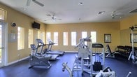 This fitness center offers a comprehensive workout environment designed to accommodate a wide range of exercise needs. The space is outfitted with a lineup of Life Fitness cardio machines, including treadmills and an elliptical trainer, positioned along a row of tall, narrow windows that allow generous natural light to fill the room. The warm yellow walls contribute to an energizing atmosphere, while the dark rubberized flooring provides a durable, safety-conscious surface throughout. On the strength training side, a multi-station cable and weight machine offers versatile resistance options alongside free weight equipment. Two ceiling fans are mounted overhead, helping to maintain comfortable airflow during workouts. A wall-mounted flat-screen television is positioned for easy viewing from the cardio area, adding an entertainment option for users. Recessed lighting supplements the natural light from the windows, ensuring the space remains well-lit at all times. The single-floor layout keeps everything accessible and organized in one open area, making it easy for users to move between cardio and strength equipment. Overall, the fitness center delivers a functional and inviting environment suited for a full-body workout routine.