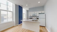 This single-floor unit showcases a thoughtfully designed open-concept layout with a clean, contemporary aesthetic. Light hardwood flooring runs throughout, complementing soft gray walls punctuated by striking navy blue accent walls that add visual depth and personality to the living and kitchen areas.

The kitchen is a functional centerpiece, anchored by a sizeable island with a speckled granite countertop and integrated sink. White cabinetry with sleek hardware lines the walls, providing ample storage, while white appliances—including a refrigerator, electric range, and built-in microwave—complete the space. Dark countertops along the perimeter offer an appealing contrast to the bright cabinetry.

Large floor-to-ceiling windows in the living area flood the space with natural light and create an airy, open feel, enhanced by elevated ceilings with recessed lighting. The living area is generously proportioned, with structural columns adding architectural character.

Bedrooms are practical and well-appointed, each featuring closet space with built-in rods—one with bifold doors and another with a walk-in configuration. The bathrooms maintain the modern palette, with one featuring a floating gray vanity, a framed mirror, and wood-look flooring, while another includes a herringbone-tiled walk-in shower.