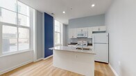 This single-floor unit showcases a thoughtfully designed open-concept layout with a clean, contemporary aesthetic. Light hardwood flooring runs throughout, complementing soft gray walls punctuated by striking navy blue accent walls that add visual depth and personality to the living and kitchen areas.

The kitchen is a functional centerpiece, anchored by a sizeable island with a speckled granite countertop and integrated sink. White cabinetry with sleek hardware lines the walls, providing ample storage, while white appliances—including a refrigerator, electric range, and built-in microwave—complete the space. Dark countertops along the perimeter offer an appealing contrast to the bright cabinetry.

Large floor-to-ceiling windows in the living area flood the space with natural light and create an airy, open feel, enhanced by elevated ceilings with recessed lighting. The living area is generously proportioned, with structural columns adding architectural character.

Bedrooms are practical and well-appointed, each featuring closet space with built-in rods—one with bifold doors and another with a walk-in configuration. The bathrooms maintain the modern palette, with one featuring a floating gray vanity, a framed mirror, and wood-look flooring, while another includes a herringbone-tiled walk-in shower.