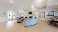 This single-story community clubhouse offers a spacious, open-concept layout designed for comfort and social interaction. The interior is finished with warm neutral tones, crown molding, wainscoting, and large-format tile flooring throughout, creating a refined yet inviting atmosphere.

The central lounge area serves as the heart of the space, anchored by a classic white-mantled fireplace with a marble surround and a wall-mounted television above. Curved upholstered sofas and accent chairs are arranged to encourage conversation, while recessed lighting and table lamps provide a warm, layered glow.

Adjacent to the lounge, a dedicated workspace area features multiple desks and chairs positioned near large windows that allow generous natural light to pour in. Decorative columns define the transitions between functional zones without interrupting the open flow of the space.

A dining area with a farmhouse-style table and upholstered chairs is situated near a glass door that opens to views of an outdoor pool and surrounding community landscape, seamlessly connecting the interior to the exterior amenity spaces.

Throughout the clubhouse, framed artwork, decorative accents, and tasteful furnishings contribute to a polished, community-oriented ambiance. The layout is thoughtfully organized to accommodate a variety of activities, from casual gatherings to more focused tasks, all within a bright and elegantly appointed setting.