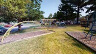 Located in Petersburg, Virginia, Ivy Gates is a residential community that combines practical living spaces with outdoor amenities in a well-established, tree-lined setting. The exterior grounds feature ample surface parking shaded by large mature trees, along with a dedicated playground area equipped with climbing structures and swing sets set on wood-chip surfacing — ideal for younger residents. Green lawns connect the various areas of the community, contributing to an open, welcoming atmosphere.

Inside, the single-floor layout is anchored by a functional kitchen outfitted with warm honey-toned wood cabinetry, white appliances including a gas range, refrigerator, dishwasher, and built-in microwave. Countertops provide generous prep space, and a pass-through opening connects the kitchen to adjacent living areas, enhancing the sense of openness. Natural light enters through blinded windows, brightening the neutral-toned interior. Durable tile-style flooring runs throughout the main living areas, offering easy maintenance.

A staircase with metal railing is visible near the entry, suggesting multi-level units may be available within the community. Overall, Ivy Gates presents a straightforward, functional residential environment suited for everyday living.
