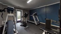Located at 2400 Milhaven Dr in Henrico, Virginia, this single-floor fitness center is a well-appointed exercise space designed to support a full range of workout routines. The room features a two-tone color scheme with bold navy blue and warm taupe walls, accented by crisp white trim and crown molding that lend the space a clean, polished appearance. A large wall-mounted mirror spans one side of the room, reflecting the full layout and amplifying the sense of space, while an inspirational wall decal reading "Everyday Is A Good Day To Work Out" adds motivational character. The fitness area is equipped with cardio machines including a treadmill and stationary bike, as well as strength training equipment and free weights. Stability balls and additional accessories round out the offerings. Durable rubber flooring throughout provides safety and comfort underfoot. Multiple windows along the exterior wall invite natural light and offer views of the surrounding greenery, creating an energizing atmosphere. A wall-mounted flat-screen television is positioned for convenient viewing during workouts. A keyed entry door with exterior access connects the space directly to the outdoor environment, enhancing ventilation and accessibility. Overhead fluorescent lighting ensures the room remains bright and functional at all hours.