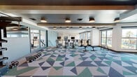 Located at 1000 North 44th Street in Phoenix, AZ, this two-story fitness center offers a comprehensive workout facility designed with both function and aesthetics in mind. The lower floor houses the primary strength and cardio zones, featuring an extensive free weights area with a full dumbbell rack, adjustable benches, and multiple cable and resistance machines. Large floor-to-ceiling windows flood the space with natural light and provide views of the surrounding outdoor amenity areas, including a pool deck with palm trees. A distinctive geometric-patterned carpet in tones of green, blue, and gray anchors the main floor, complementing the dark ceiling with warm drum pendant lighting and wood-accented architectural beams. A central staircase with modern cable railings connects to the upper floor, which features a dedicated cycling and rowing studio with hard flooring, wall-mounted TVs, and expansive windows with outdoor views. An adjoining functional training studio on the upper level offers light wood flooring, a textured tile accent wall, and access to a medicine ball and kettlebell rack. The lower level also includes a strength-focused room with rubber flooring, a multi-cable functional trainer, barbell storage, and direct outdoor access. Throughout both floors, full-length mirrors maximize the sense of space and support training visibility.
