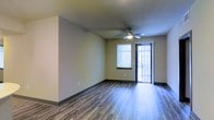 This single-floor unit offers a clean, modern layout designed for comfortable everyday living. The main living area is a standout feature, showcasing durable wood-look plank flooring that runs throughout, complemented by dark baseboards that add a polished, contrasting finish. Neutral-toned walls provide a versatile backdrop, making the space easy to personalize.

A ceiling fan with an integrated light fixture is centrally positioned overhead, ensuring both comfortable airflow and ambient illumination throughout the room. Natural light pours in through a horizontal window fitted with blinds, brightening the interior and creating an inviting atmosphere.

A sliding glass door with vertical blinds opens directly to an outdoor area, offering a seamless connection between the interior living space and the outside. This feature not only enhances ventilation options but also adds an additional source of natural light, contributing to the airy feel of the room.

The overall design of this space is straightforward and functional, with a well-considered layout that maximizes usability. The combination of contemporary flooring, neutral finishes, and access to outdoor space makes this a practical and welcoming single-floor unit suited to a variety of lifestyles.