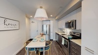 This contemporary single-floor residence offers a thoughtfully designed open-concept layout that seamlessly connects the kitchen, dining, and living areas. The kitchen is equipped with stainless steel appliances, including a French door refrigerator, electric range, built-in microwave, and dishwasher. Sleek dark lower cabinetry is paired with high-gloss white upper cabinets and a subway tile backsplash. A white dining island with stylish gold-accented seating sits beneath a striking oversized pendant light, creating a functional and visually appealing dining space. Rich dark hardwood-style flooring flows throughout the main living areas, adding warmth and continuity. The living room is bathed in natural light from large sliding glass doors that open to an outdoor balcony, offering views of the surrounding neighborhood. The bedroom features a walk-in closet with built-in shelving, a window with natural light, and the same hardwood-style flooring. The bathroom presents a clean, modern aesthetic with large-format tile flooring, a walk-in shower with a handheld fixture, a floating vanity with an undermount sink, and an illuminated mirror. The overall ambiance is bright, contemporary, and move-in ready.