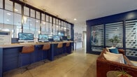 This thoughtfully designed single-floor communal facility blends modern industrial aesthetics with warm, inviting elements throughout its open layout. Polished concrete floors unify the entire space, complementing bold navy blue accent walls and a mix of textures that give the interior a distinctive, contemporary character.

A dedicated business center features a row of iMac workstations set along a reclaimed wood countertop mounted against a deep blue shiplap wall, framed by an elegant steel-and-glass partition with pendant glass bulb lighting overhead. Leather and metal bar stools provide comfortable seating for focused work sessions.

The lounge areas offer multiple seating arrangements, including leather sofas, upholstered armchairs in mustard yellow and deep blue, and low coffee tables arranged on patterned area rugs. An arched architectural feature wall displays an inspirational quote, adding a creative focal point, while a striking stone fireplace anchors another sitting nook. Large windows throughout flood the space with natural light and provide views to the outdoors.

A café-style bar area is highlighted by an eye-catching mosaic tile backdrop, navy cabinetry, gold bar stools, and colorful pendant lighting in aqua blue. Transparent acrylic chairs and round tables complete this social gathering space. Framed artwork and curated wall graphics are displayed throughout, contributing to the space's vibrant, community-oriented atmosphere.