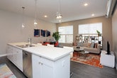 This single-floor unit showcases a contemporary design with a neutral gray palette, dark hardwood-style flooring, and exposed ductwork that adds an industrial accent. The open living area is anchored by large windows with horizontal blinds that invite natural light and street views, complemented by a modular shelving unit and a vibrant orange area rug that adds warmth to the space.

The kitchen is well-appointed with two-toned cabinetry—matte dark upper cabinets paired with clean white lower cabinets—set against a tile backsplash. Quartz countertops extend across the workspace and onto a central island with bar seating, illuminated by a geometric pendant light. Stainless steel appliances complete the functional layout.

The primary bedroom features elevated ceilings with exposed ductwork, an en-suite bathroom with a white vanity and walk-in closet area. A second bedroom offers a quieter, more compact layout. A second bathroom includes a white marble-top vanity and a bold graffiti-style shower curtain as a striking design statement.

A separate dining or flex room provides a defined space for work or entertaining, finished with tile flooring and recessed lighting.