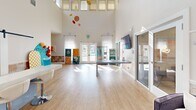 Located at 700 Berkeley Place Circle in Clemson, South Carolina, this single-floor leasing and community space for Clemson Edge offers a vibrant and welcoming environment designed to impress. The open-plan layout is anchored by dramatically soaring ceilings that amplify the sense of space, while a continuous row of upper clerestory windows floods the interior with abundant natural light. Light wood-look flooring runs throughout, lending warmth and continuity to the space. A prominent front reception counter with modern bar-height seating serves as the central greeting point, complemented by a sliding barn door that adds an on-trend architectural detail. Glass-paneled doors provide a view into an adjacent private office or consultation room, maintaining an open and transparent feel. A wall-mounted flat-screen display, a central high-top table, and a comfortable seating area with upholstered chairs create distinct functional zones within the single room. Sheer white curtains frame the large front-facing doors, which open to outdoor views and allow natural light to pour in. The overall ambiance is bright, energetic, and modern, reflecting the youthful and dynamic character of the Clemson Edge community.
