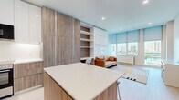 Located at 2100 N Central Rd in Fort Lee, NJ, this thoughtfully designed studio offers a compact yet highly functional layout across a single floor. The open-plan living area is bathed in natural light through large floor-to-ceiling windows fitted with roller shades, revealing sweeping views of the surrounding cityscape and greenery. Light hardwood flooring runs throughout, lending a clean and cohesive aesthetic to the space.

The kitchen is a standout feature, showcasing two-tone cabinetry that combines warm wood-grain lower cabinets with crisp white upper units. A central island with a white quartz countertop and bar seating provides additional prep space and casual dining. Stainless steel appliances, including a range and microwave, complete the modern setup.

A built-in wall unit adjacent to the kitchen integrates floor-to-ceiling storage panels alongside open shelving and a compact desk area, maximizing space efficiency. The living area comfortably accommodates a sofa and console table, with the generous windows serving as a natural focal point.

The bathroom is elegantly finished with marble-veined wall tiles, large-format floor tiles, a frameless glass shower enclosure, and a floating wood-finish vanity topped with a white basin — all accented by polished chrome fixtures for a refined, contemporary feel.