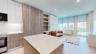 Located at 2100 N Central Rd in Fort Lee, NJ, this thoughtfully designed studio offers a compact yet highly functional layout across a single floor. The open-plan living area is bathed in natural light through large floor-to-ceiling windows fitted with roller shades, revealing sweeping views of the surrounding cityscape and greenery. Light hardwood flooring runs throughout, lending a clean and cohesive aesthetic to the space.

The kitchen is a standout feature, showcasing two-tone cabinetry that combines warm wood-grain lower cabinets with crisp white upper units. A central island with a white quartz countertop and bar seating provides additional prep space and casual dining. Stainless steel appliances, including a range and microwave, complete the modern setup.

A built-in wall unit adjacent to the kitchen integrates floor-to-ceiling storage panels alongside open shelving and a compact desk area, maximizing space efficiency. The living area comfortably accommodates a sofa and console table, with the generous windows serving as a natural focal point.

The bathroom is elegantly finished with marble-veined wall tiles, large-format floor tiles, a frameless glass shower enclosure, and a floating wood-finish vanity topped with a white basin — all accented by polished chrome fixtures for a refined, contemporary feel.