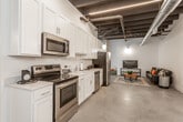This single-floor loft showcases a striking industrial aesthetic defined by exposed black structural beams, ductwork, and polished concrete floors throughout. The open-concept design seamlessly connects the kitchen and living areas, creating a spacious and versatile environment. The fully equipped kitchen features crisp white shaker cabinetry extending to the ceiling, stainless steel appliances including a range, microwave, dishwasher, and refrigerator, and a clean white countertop with an undermount sink. Linear LED lighting runs along the dark ceiling, providing sharp, modern illumination. The living area offers a comfortable and relaxed atmosphere, anchored by a leather sofa with colorful accent pillows, a rustic coffee table, and a media console. The bedroom is generously sized with soaring ceilings accented by exposed wooden beams, offering a calm and open feel. An expansive walk-in closet provides built-in shelving and multiple hanging rods for ample storage. The bathroom is clean and functional, featuring a walk-in shower, toilet, and vanity with polished concrete flooring continuing the cohesive design language. An additional unfinished room presents flexible potential for a variety of uses. Together, these elements create a cohesive, contemporary industrial living space.