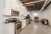 This single-floor loft showcases a striking industrial aesthetic defined by exposed black structural beams, ductwork, and polished concrete floors throughout. The open-concept design seamlessly connects the kitchen and living areas, creating a spacious and versatile environment. The fully equipped kitchen features crisp white shaker cabinetry extending to the ceiling, stainless steel appliances including a range, microwave, dishwasher, and refrigerator, and a clean white countertop with an undermount sink. Linear LED lighting runs along the dark ceiling, providing sharp, modern illumination. The living area offers a comfortable and relaxed atmosphere, anchored by a leather sofa with colorful accent pillows, a rustic coffee table, and a media console. The bedroom is generously sized with soaring ceilings accented by exposed wooden beams, offering a calm and open feel. An expansive walk-in closet provides built-in shelving and multiple hanging rods for ample storage. The bathroom is clean and functional, featuring a walk-in shower, toilet, and vanity with polished concrete flooring continuing the cohesive design language. An additional unfinished room presents flexible potential for a variety of uses. Together, these elements create a cohesive, contemporary industrial living space.