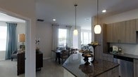 This single-floor residence showcases an open-concept design that integrates the kitchen, dining, and living areas into one cohesive, flowing space. The kitchen is appointed with rich dark granite countertops that provide a striking contrast against the light-toned cabinetry, complemented by pendant lighting that hangs elegantly over the counter's edge, adding both function and style. Recessed lighting throughout ensures consistent, well-distributed illumination across the space.

The dining area accommodates a dark wood table paired with matching lattice-back chairs, anchored beneath additional pendant lighting that defines the zone within the open layout. Patterned curtains frame the windows, allowing natural light to filter in while contributing a soft, decorative touch to the neutral color palette of white walls and light hardwood-style flooring.

A sideboard with a ribbed front panel adds storage and character along one wall, while a decorative pedestal table near the kitchen bar adds a refined accent. The living area is partially visible beyond the dining space, reinforcing the home's spacious, connected ambiance suited for everyday living and entertaining.