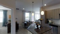 This single-floor residence showcases an open-concept design that integrates the kitchen, dining, and living areas into one cohesive, flowing space. The kitchen is appointed with rich dark granite countertops that provide a striking contrast against the light-toned cabinetry, complemented by pendant lighting that hangs elegantly over the counter's edge, adding both function and style. Recessed lighting throughout ensures consistent, well-distributed illumination across the space.

The dining area accommodates a dark wood table paired with matching lattice-back chairs, anchored beneath additional pendant lighting that defines the zone within the open layout. Patterned curtains frame the windows, allowing natural light to filter in while contributing a soft, decorative touch to the neutral color palette of white walls and light hardwood-style flooring.

A sideboard with a ribbed front panel adds storage and character along one wall, while a decorative pedestal table near the kitchen bar adds a refined accent. The living area is partially visible beyond the dining space, reinforcing the home's spacious, connected ambiance suited for everyday living and entertaining.