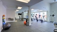 This fitness center is designed to support a comprehensive workout experience within a bright, open layout. The space is finished with durable rubber flooring throughout, providing both safety and comfort during exercise. Large windows along one wall allow ample natural light to fill the room, creating an energizing atmosphere while offering views of the surrounding exterior. Recessed panel lighting in the ceiling supplements natural light, ensuring the space remains well-illuminated at all times. Structural columns divide the floor plan into distinct zones without compromising the open feel of the room. The cardio area is lined with a row of elliptical trainers, stationary bikes, and treadmills, each equipped with individual monitor screens. A dedicated strength training zone houses a multi-use cable machine and weight bench, accommodating a variety of resistance exercises. A wall-mounted flat-screen television and mirrored surfaces enhance the workout environment, while a water station and exercise ball are conveniently available. The neutral white walls and grey ceiling contribute to the clean, modern aesthetic of the space overall.